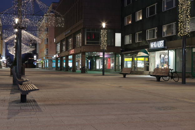 Eine nächtliche Straßenansicht mit Straßenlaternen, Weihnachtsbeleuchtung, Bänken, Pfosten, Fahrrädern, Gebäuden mit Fenstern, Namensschildern und einem Sternenhimmel.