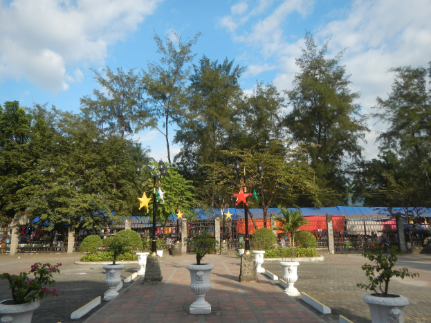 Ein Park mit Topfpflanzen und Bäumen, umgeben von Straßenlaternen, Dekorationen und Grills unter einem bewölkten Himmel.