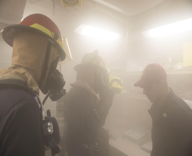 Feuerwehrleute mit Helmen und Rauch aus dem Mund, einer hält eine Kamera, während einer Übung mit Hintergrundbeleuchtung und Equipment.