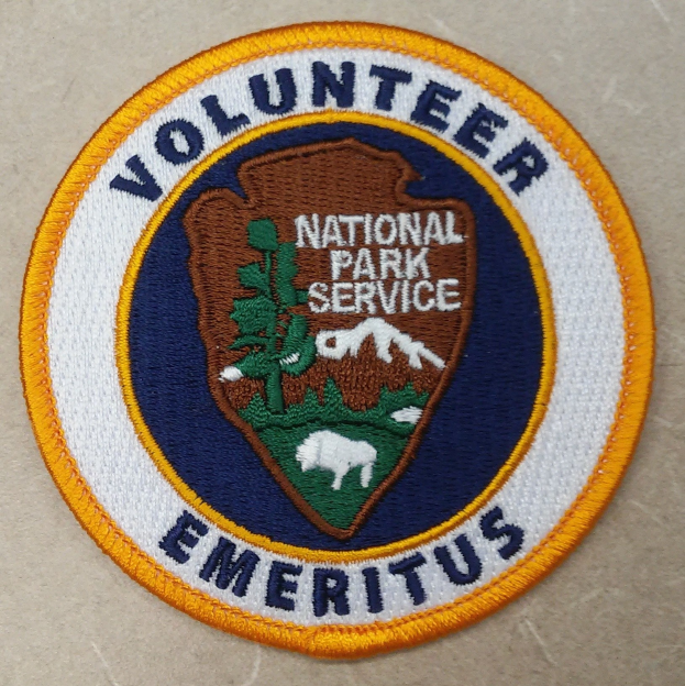 Runder Patch mit blauem Rand und weißem Hintergrund, mittig die schwarze Schrift "Volunteer National Park Service Emeritus" in einem gelben Kreis.