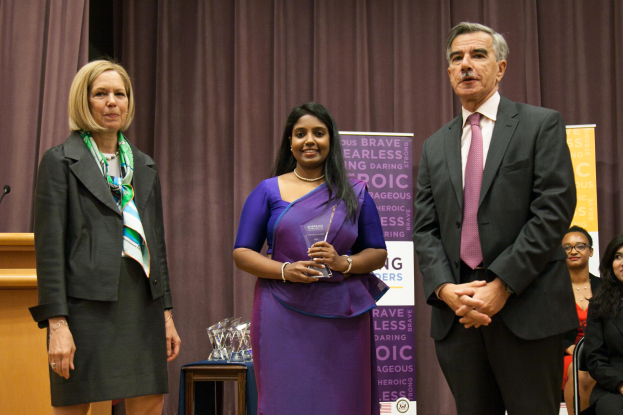 Zwei Frauen und ein Mann stehen auf einer Buhne mit einem braunen Vorhang dahinter; die Frau in der Mitte halt einen Preis, und mehrere Preise sind auf einem Tisch vor ihnen.