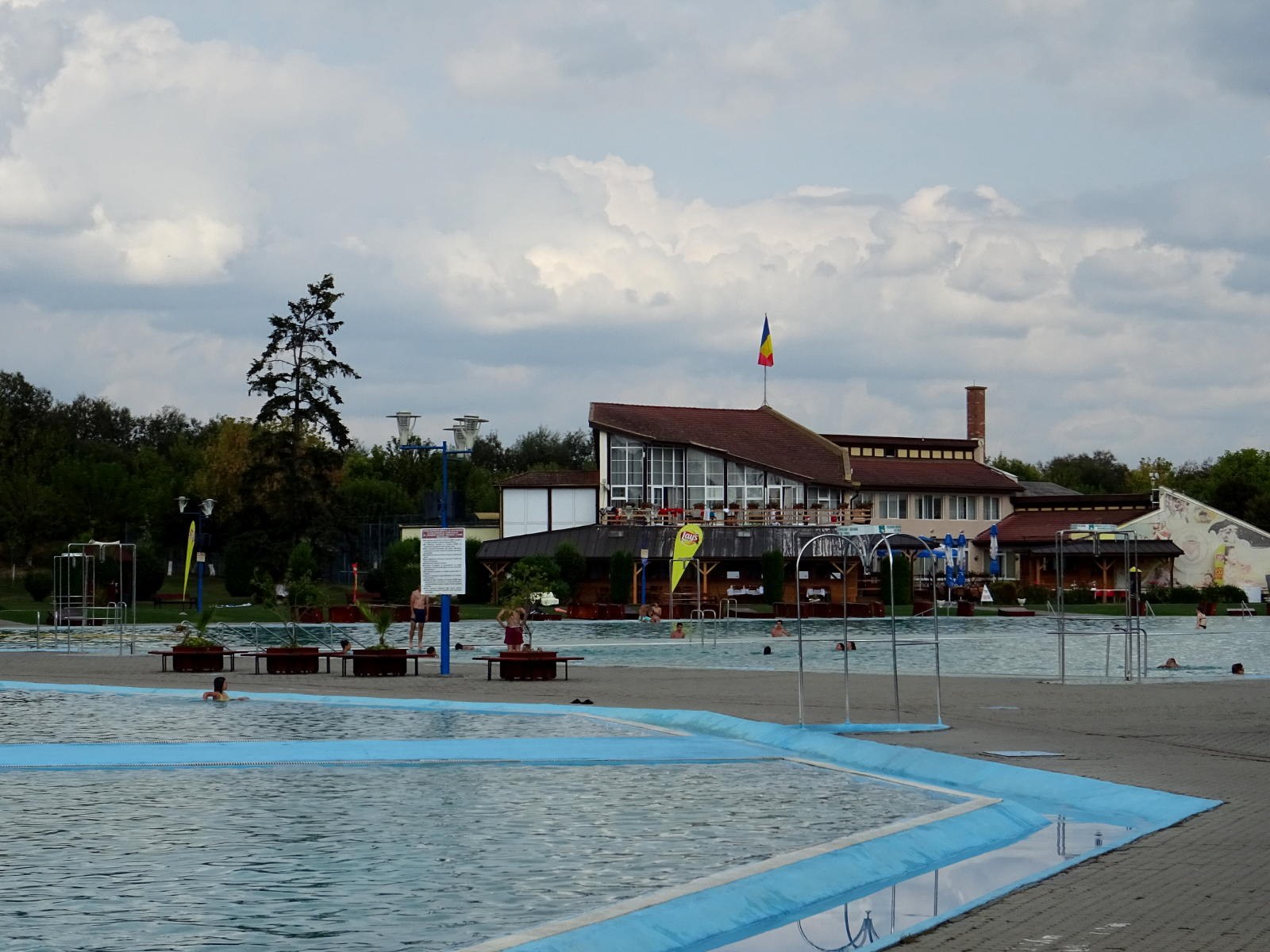 Ein großes Schwimmbecken mit Menschen darin, umgeben von Pfählen, Bänken, Topfpflanzen, einem Schild, einem Fahnenmast mit einer Fahne, einem Gebäude mit Fenstern, Straßenlaternen, einer Baumgruppe und einem bewölkten Himmel.