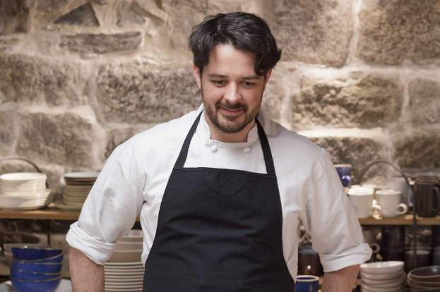 Ein Koch in einer weißen Schürze und einer schwarzen Schürze steht vor einem Regal mit Tellern, Schüsseln und Tassen an einer Wand.