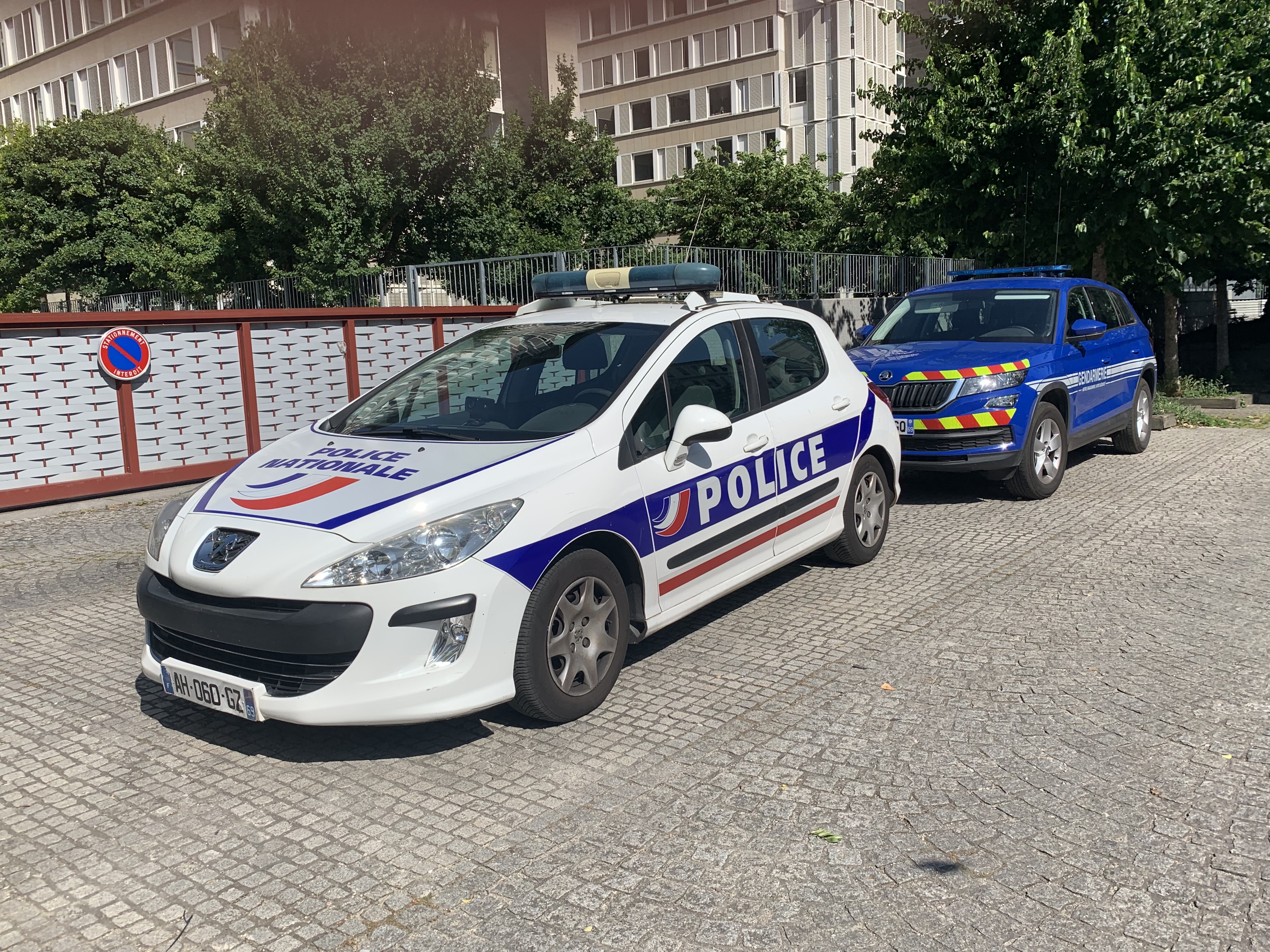 Zwei Polizeiwagen, ein Peugeot 308 und ein Citroën C3, nebeneinander in einem Wohngebiet mit Bäumen und Gebäuden im Hintergrund geparkt, und ein Schild an einem Pfahl links daneben.