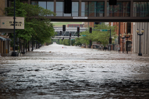 Überschwemmte Stadtstraße mit Wasser, das die Straße, Pfosten, Laternen, Schilder, Verkehrszeichen, Bäume, Gebäude und eine ferne Brücke bedeckt.