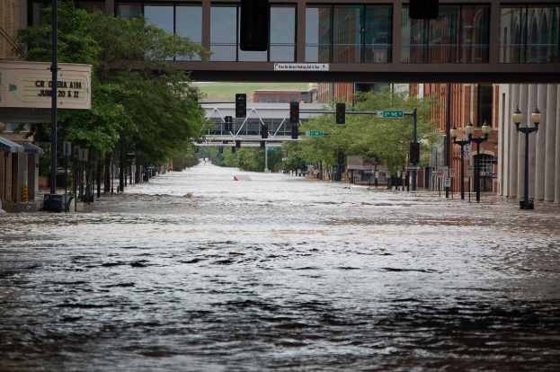 Überschwemmte Stadtstraße mit Wasser, das die Straße, Pfosten, Laternen, Schilder, Verkehrszeichen, Bäume, Gebäude und eine ferne Brücke bedeckt.