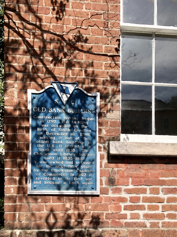 Altes Bankgebäude mit blauer Tafel an der Seite, Fenster rechts und Baum links; die Tafel enthält Text.