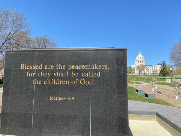 Ein Gedenkstein mit der Inschrift "Selig sind die Friedenstifter, denn sie werden Kinder Gottes genannt" umgeben von Bäumen, Pflanzen, Gras, Laternenmasten und einem Gebäude unter einem klaren blauen Himmel.