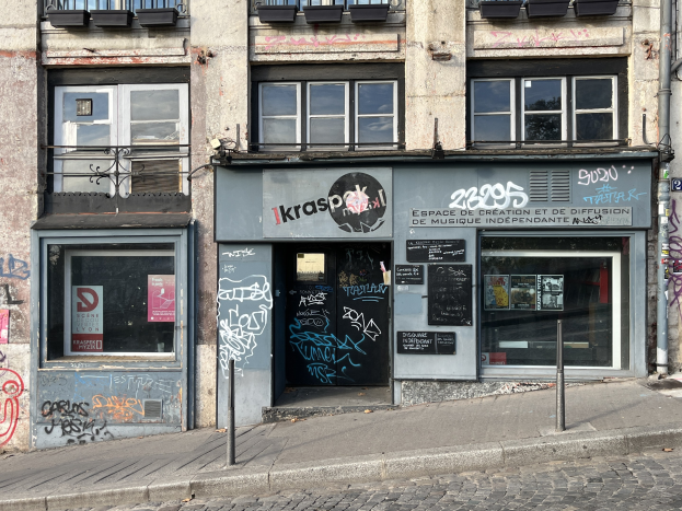 Ein Gebäude mit Fenstern, Geländern, Türen, Texttafeln, Plakaten, Pfählen und Graffiti an seiner Seite, das über einer Straße liegt.