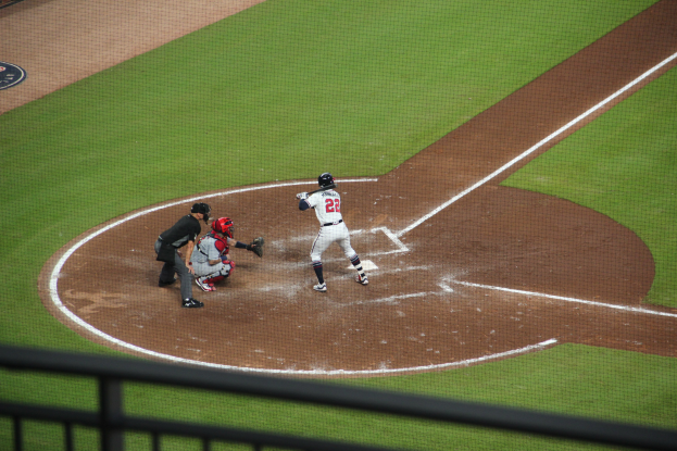 Ein Baseball-Spieler in einem Sportdress schwingt einen Schläger auf einem grünen Feld, mit einem Geländer am unteren Bildrand.