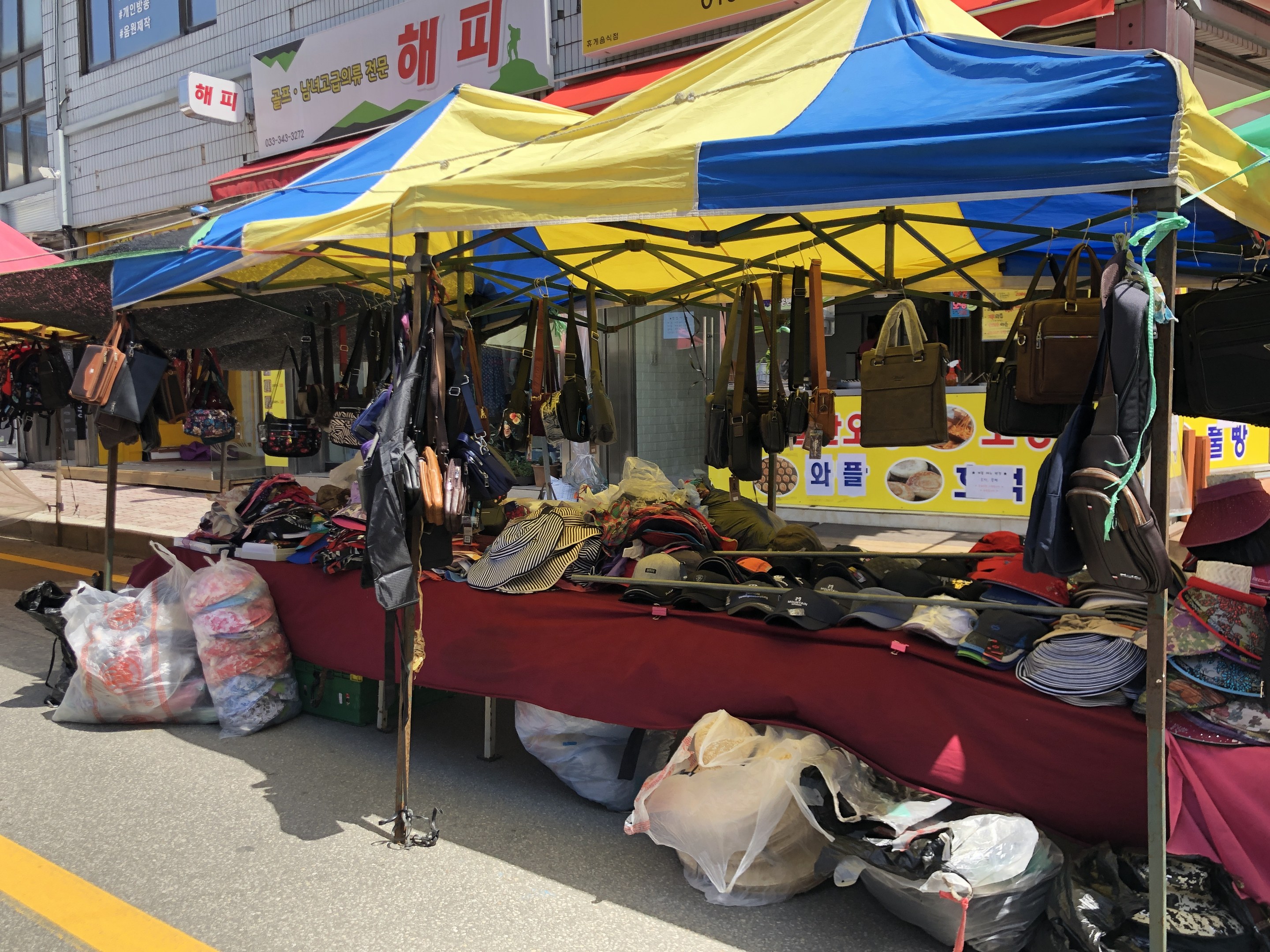 Ein Markt mit Zelten, die verschiedene Gegenstände wie Taschen und Hüte präsentieren, vor Häusern mit Fenstern und Texttafeln.