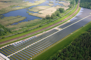 Luftaufnahme einer Solaranlage mit Panelen in einem Feld, umgeben von Bäumen, Gras, Pflanzen und Wasser, mit einem Zug auf einer nahen Eisenbahnstrecke.