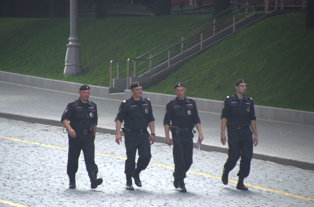 Vier Polizisten in Uniform gehen die Straße entlang und halten Papiere in der Hand, mit einem Gebäude, Bäumen und einem Pfahl im Hintergrund.