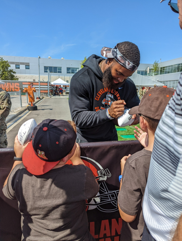 Cleveland Browns Wide Receiver Odell Beckham Jr. signiert Autogramme für Fans auf dem Trainingsgelände, umgeben von Menschen mit Mützen, einem Banner, Gebäuden, Bäumen und Zelten im Hintergrund unter einem bewölkten Himmel.