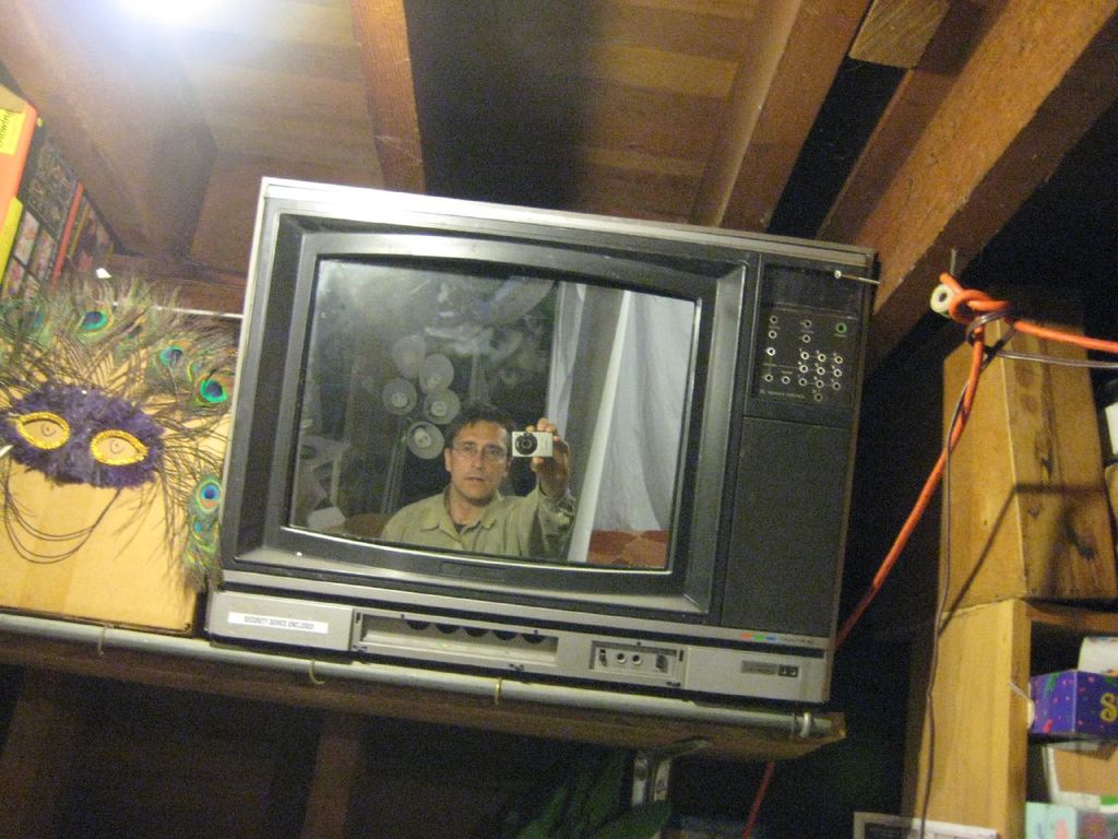 Ein Fernsehbildschirm mit einer Person umgeben von Pfauenfedern, Büchern und Schachteln.