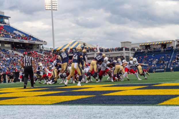 Fußballspieler in Helmen und Sportkleidung konkurrieren auf einem Feld mit Zuschauern, Stadionstrukturen und Text unten.