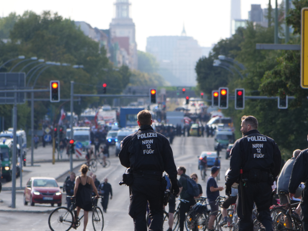 A group of police officers riding bicycles down a tree-lined street with buildings and a clear blue sky in the background.