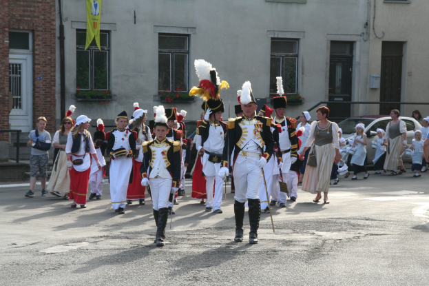 Eine Gruppe von Menschen, die auf der Straße in einer Parade gehen, einige in Kostümen und mit Stöcken, mit Gebäuden, Pflanzen, einer Flagge und Fahrzeugen im Hintergrund.