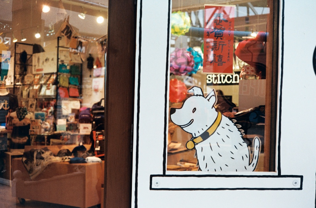 Eine künstlerische Darstellung eines Hundes mit begleitendem Text auf der rechten Seite und links ein Raum voller verschiedener Gegenstände wie Lampen, Flaggen, Regalen, Sofas, Kleidung, Dekorationen, Menschen und Stühlen.