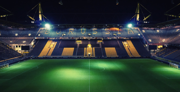 Ein Fußballfeld bei Nacht, beleuchtet von hellen Stadionlichtern, mit einigen Menschen auf den Tribünen und grünem Rasen auf dem Boden.