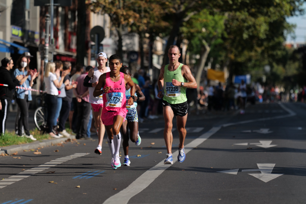 Gruppe von Menschen, die bei einem Marathon auf einer Stadtstraße laufen, mit Zuschauern und unscharfen Hintergrund-Elementen wie Bäumen, Gebäuden und einem Fahrrad.