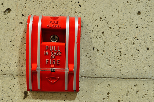 Roter zylindrischer Feueralarmkasten mit glänzender Oberfläche, an einem Gebäude montiert, mit einem oberen Griff, einem zentralen Schlüsselloch und der auffälligen schwarzen Aufschrift "Feueralarm".