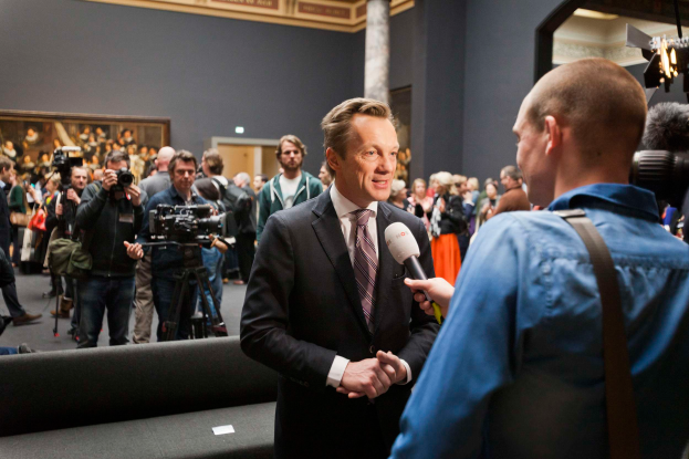 Ein Mann im Anzug und Krawatte wird von einem Reporter interviewt, vor einem Publikum mit einigen Kameras, einer Couch im Vordergrund und einer Wand mit einem Foto, Lampen und einer Säule im Hintergrund.