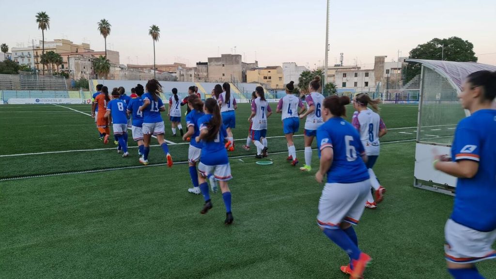 Eine Gruppe von Frauen, die auf einem Feld mit Gebäuden, Bäumen und Pfählen umgeben von einem klaren blauen Himmel Fußball spielen, mit einem Torpfosten und Netz auf der rechten Seite.
