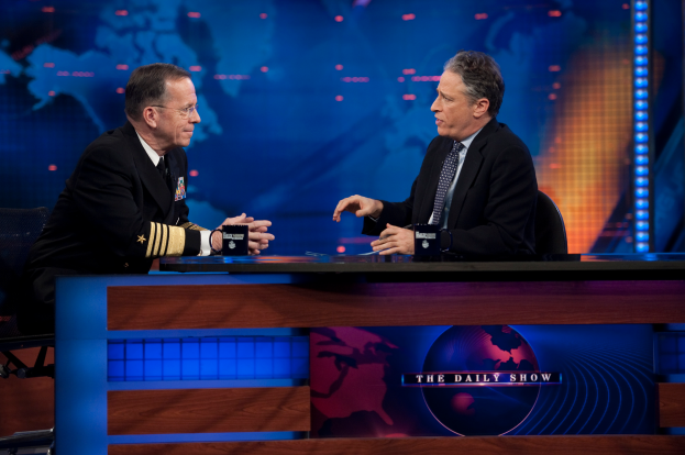 Zwei Männer in schwarzen Blazern, Hemden und Krawatten sitzen an einem Tisch mit Mikrofonen dazwischen und unterhalten sich vor einem TV-Bildschirm und einem blauen Hintergrund.