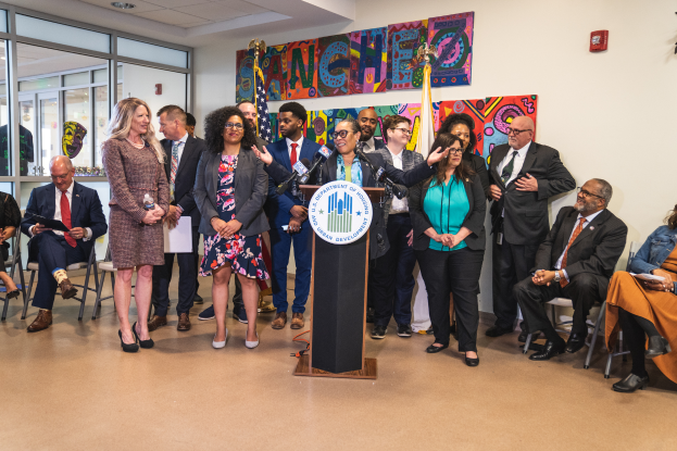Eine Gruppe von Menschen, die in einem Raum stehen und sitzen, mit einem Podium, einem Mikrofon und einer Tafel mit der Aufschrift "Sancho-Stadtrat tritt dem Stadtrat bei", mit Flaggen, einem Feuerlöscher und Deckenleuchten an der Rückwand.