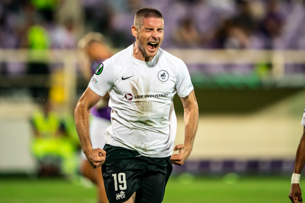 Ein Mann in weißem Trikot und schwarzen Shorts rennt auf einem Fußballfeld, feiert ein Tor, umgeben von einer Gruppe von Menschen, mit einem unscharfen Hintergrund.