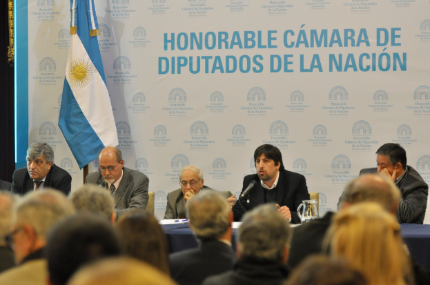 Eine Gruppe von Menschen sitzt in Reihen von Stühlen, mit einem Tisch vor der ersten Reihe, auf dem ein Krug, Papiere und ein Stift stehen. Eine Person in der Mitte spricht in ein Mikrofon, hinter ihr eine weiße und blaue Flagge und ein weißer und blauer Hintergrund mit der Aufschrift "ehrenwerte kamara de" im Hintergrund.