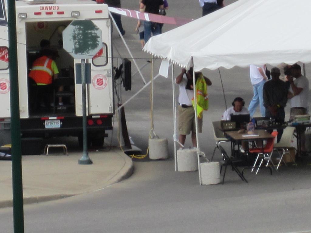Ein Krankenwagen steht links von einer Stra√üe, w√§hrend auf der rechten Seite ein Zelt viele Menschen sch√ľtzt, mit einem Tisch und Stuhl in der N√§he.