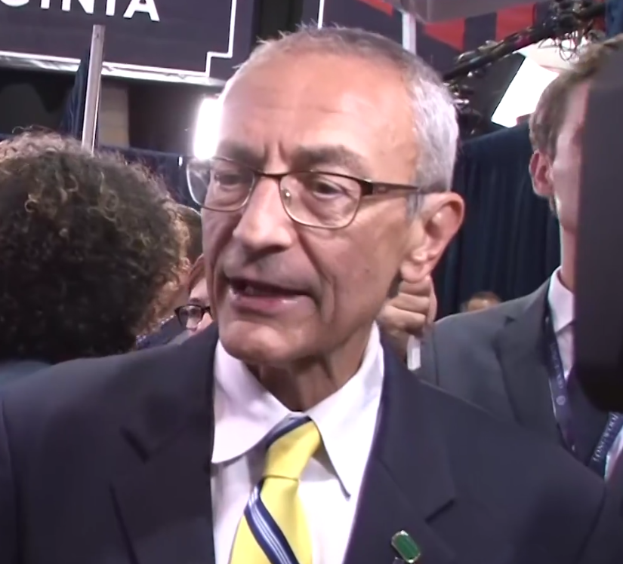 Ein Mann im Anzug und Krawatte mit Brille spricht zu Reportern, umgeben von einer Gruppe von Menschen, mit einem Mikrofonständer vor ihm und einer Tafel mit Text im Hintergrund.