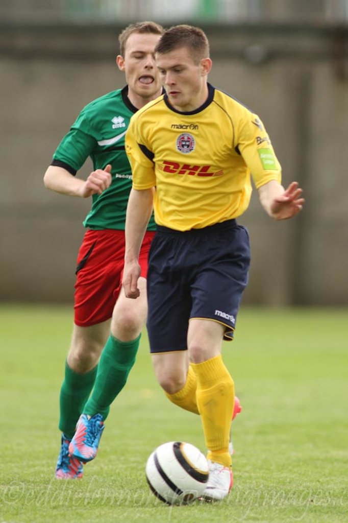 Zwei Männer spielen Füßball auf einem grünen Feld mit einem Ball, mit unscharfem Hintergrund und Text unten.