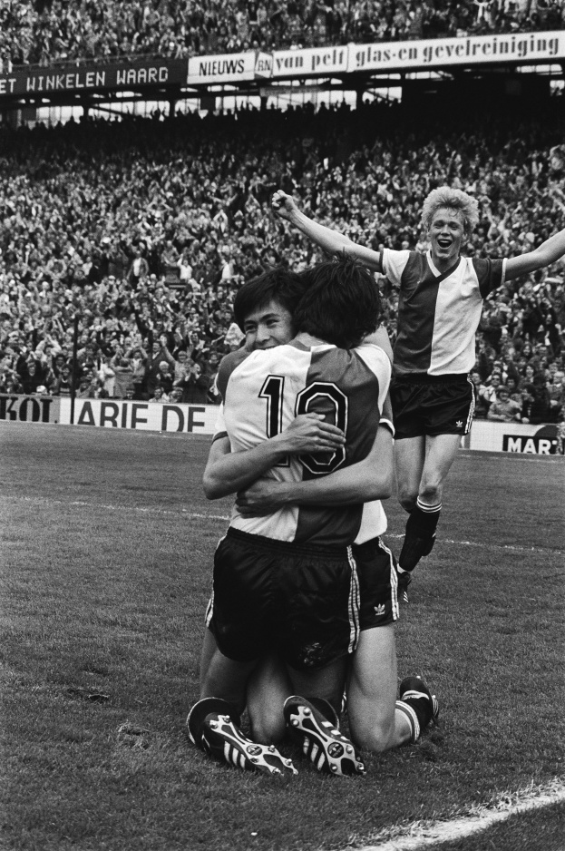 Drei Fußballer umarmen sich auf dem Boden und feiern ein Tor, mit jubelnder Menge und schwenkenden Fahnen im Hintergrund, dargestellt in Schwarz-Weiß.