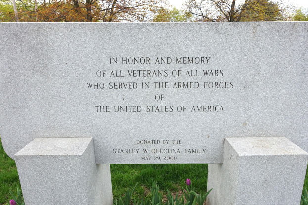Gedenkstein mit einer Widmung für US-Streitkräfte-Veteranen, umgeben von Pflanzen, Blumen, Bäumen und einem bewölkten Himmel.
