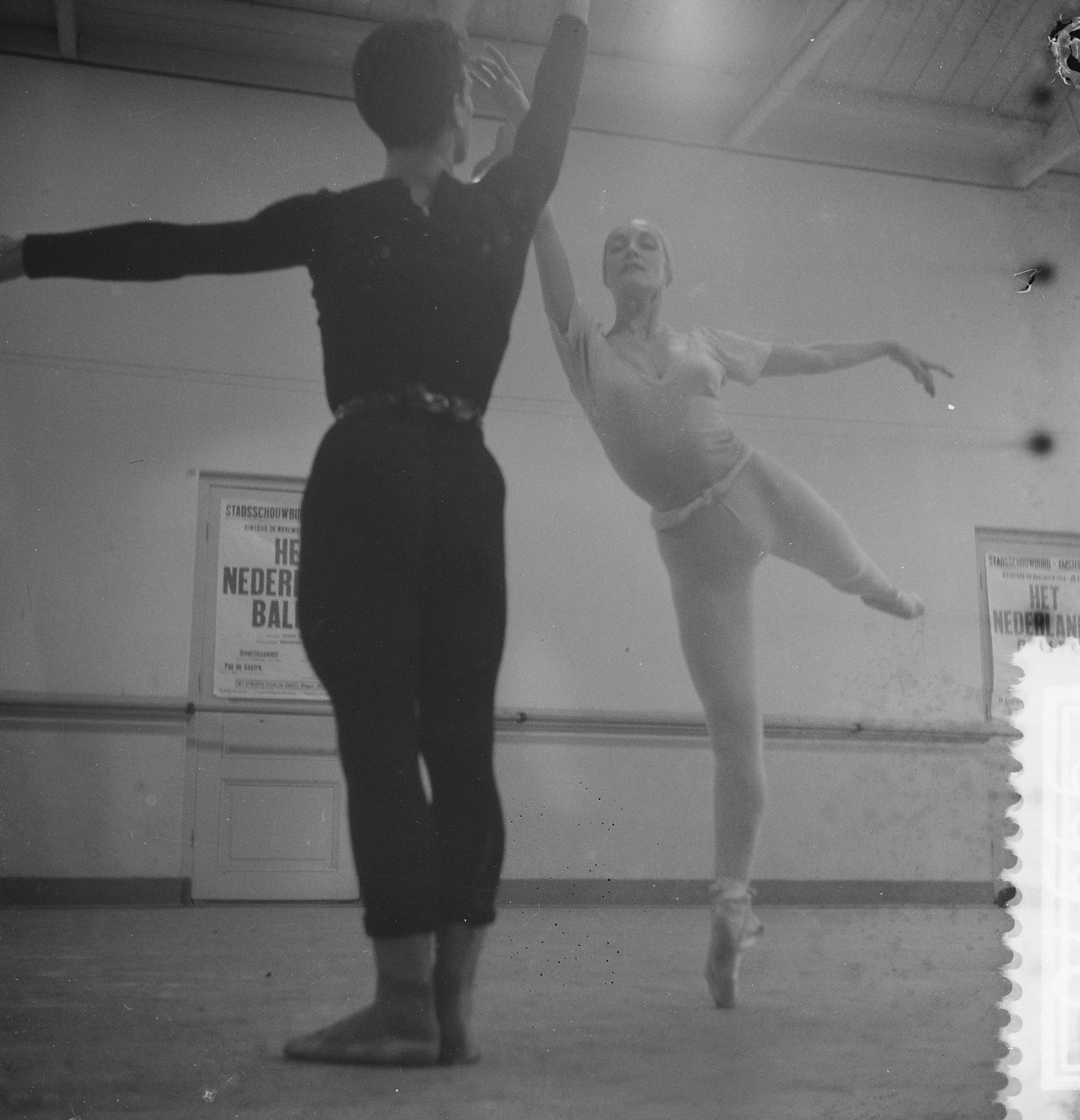 Ein Schwarz-Weiß-Foto von einem Mann und einer Frau, die in einem Studio tanzen, wobei die Frau in der Luft ist und eine Wand mit Postern im Hintergrund zu sehen ist, die von oben beleuchtet wird.