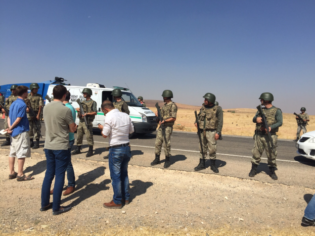 Eine Gruppe von Männern, einige mit Waffen, steht auf einem Schotterweg mit Fahrzeugen und einer Person mit einer Kamera, mit Bäumen und einem klaren blauen Himmel im Hintergrund.