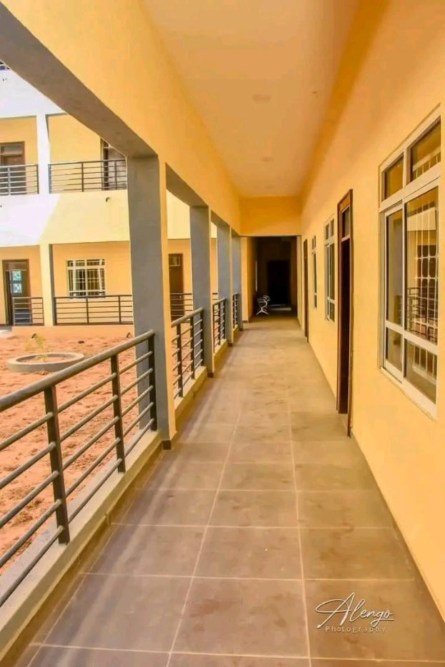 Ein mehrgeschossiges Gebäude mit Fenstern, Türen, Säulen und Geländern zeigt einen langen gefliesten Flur mit einem Balkon, umgeben von anderen Gebäuden und einem klaren blauen Himmel, mit Miettext unten.