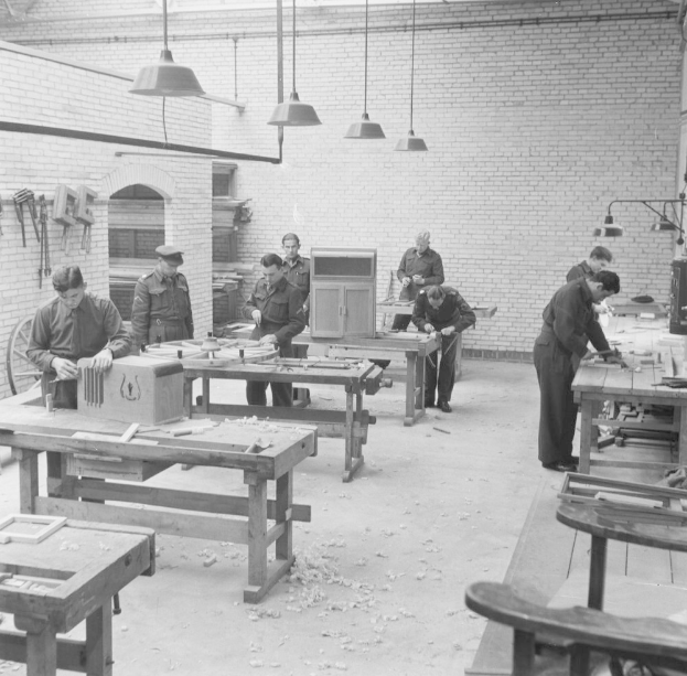 Ein Schwarz-Weiß-Bild einer Gruppe von Männern, die in einer Fabrik arbeiten, umgeben von Tischen, Stühlen und verschiedenen Werkzeugen.