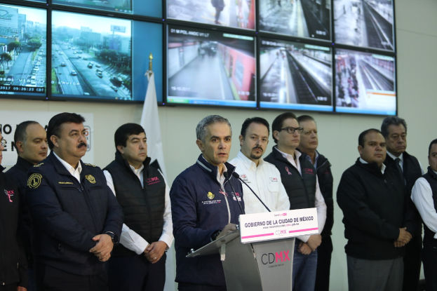 Eine Gruppe von Männern, die vor einem Podium mit einem Mikrofon und einer Tafel mit der Aufschrift "Mexikanische Zoll- und Grenzschutzbehörde" stehen, mit Fernsehgeräten und einer Flagge an der Wand dahinter.