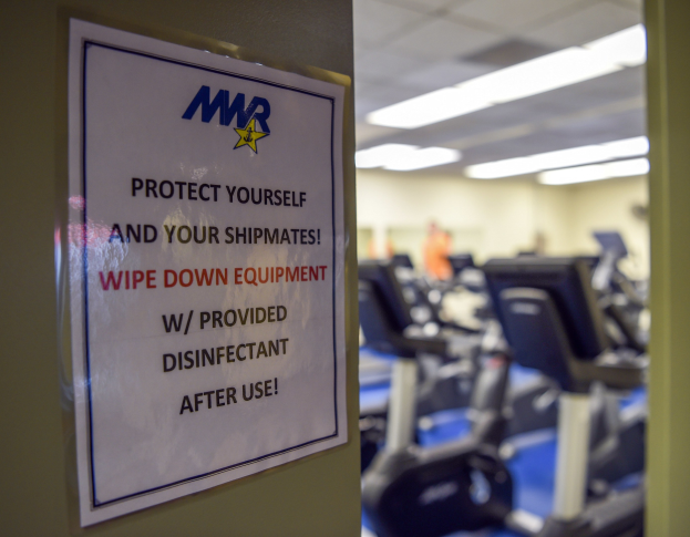 Ein Türschild mit der Aufschrift 'Schützen Sie sich und Ihre Schiffskameraden' mit Laufbändern und Deckenleuchten im Hintergrund.