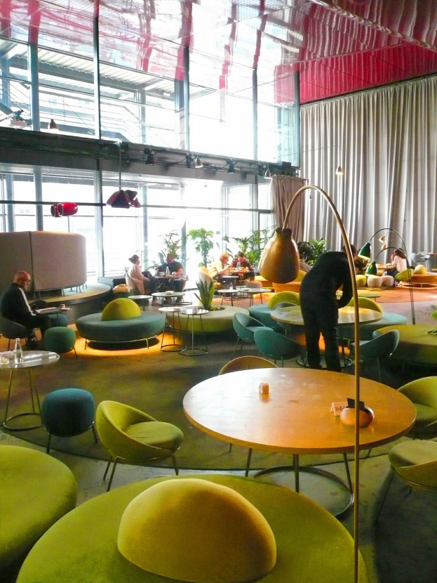 Ein geräumiger Hotellounge mit grünen Stühlen, Tischen, Sofas, stehenden Menschen, Topfpflanzen, einer Stehlampe, Vorhängen und Deckenleuchten.