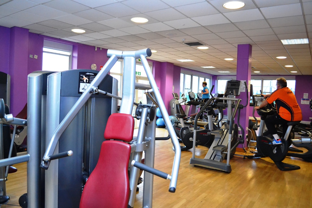 Ein Fitnessstudio mit verschiedenen Geräten und einigen Menschen beim Training, mit einer Decke und Lampen oben im Bild.