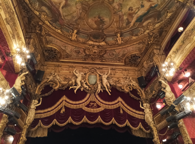 Theaterinnenraum mit roter Gardine, Kronleuchtern, Deckenmalerei und Skulpturen, mit einer Bühne und einigen Menschen davor.