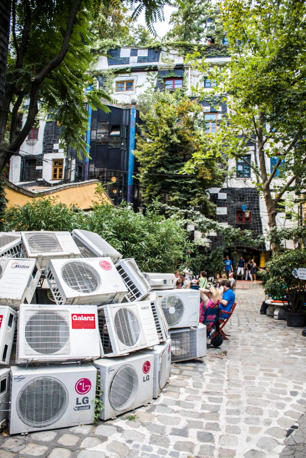 Luftkühler am Boden links unten, eine Straße rechts daneben, Menschen auf Stühlen neben der Straße sitzend, mit Gebäuden, Bäumen und Pflanzen darüber.