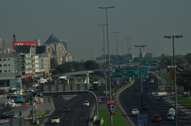 Luftaufnahme einer Stadt mit Gebäuden, Türmen, Fahrzeugen, Werbetafeln, Schildern, Straßen, Bäumen, Straßenlaternen, Menschen, Absperrgittern und Informationsschildern unter einem klaren Himmel.