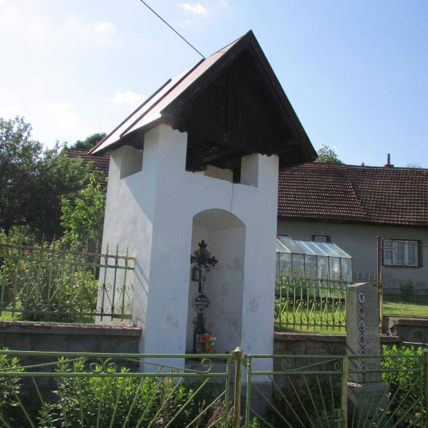 Kleine weiße Kirche mit einem Kreuz darauf, umgeben von einem Metallzaun, Pflanzen, Gras, einem Weg, nahen Häusern, Pfählen, Drähten und einem bewölkten Himmel, identifiziert als die Kirche des Heiligen Grabes.