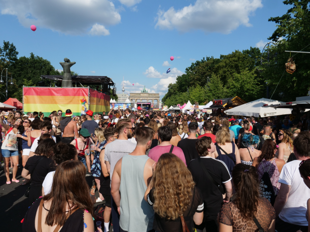 Eine große Menschenmenge, die eine Straße mit Zelten, Bäumen, Pfählen, Lichtern und einer Statue gesäumt, mit Gebäuden und einem bewölkten Himmel mit Ballons im Hintergrund beim Berlin Pride Festival.